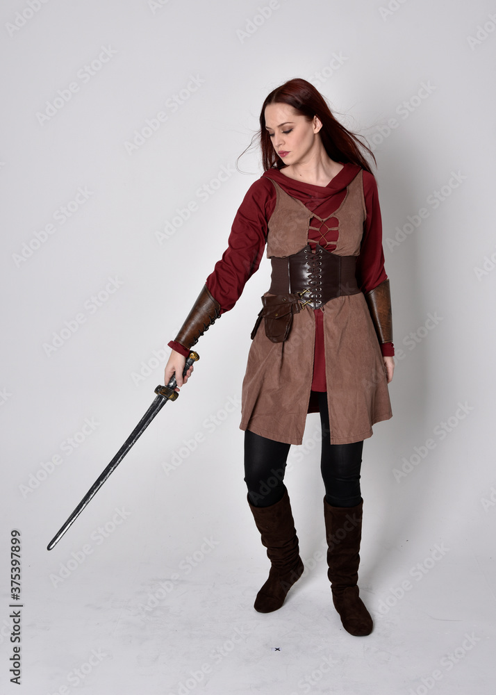 Full length portrait of girl wearing medieval costume. Standing pose holding a sword, isolated against a grey studio background. Full length portrait of girl wearing medieval costume. Standing pose holding a sword, isolated against a grey studio background.