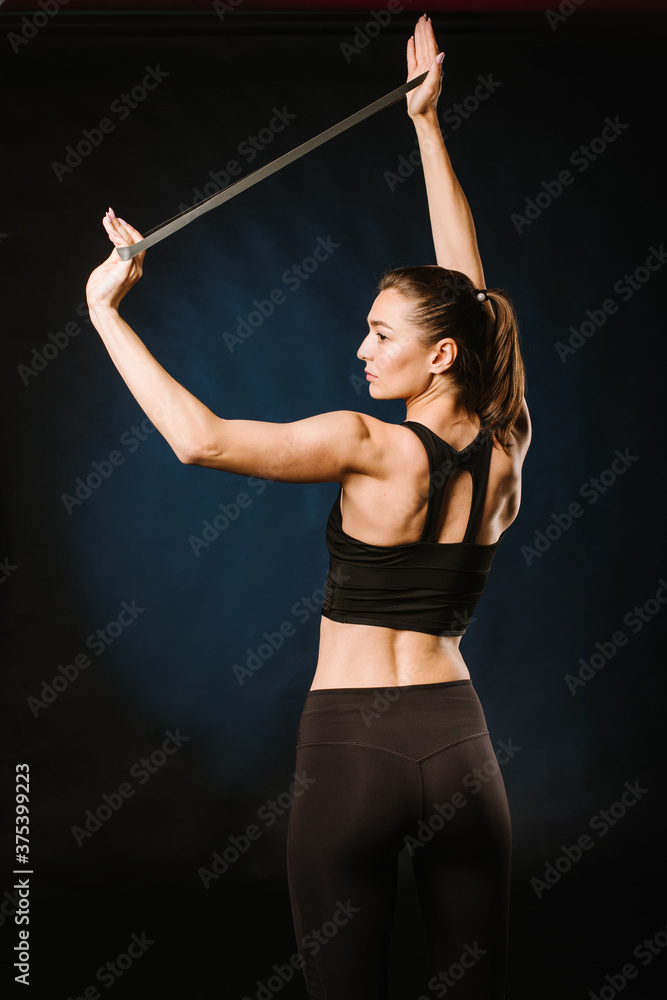 Young, slim girl doing exercises with rubber expander on black background.