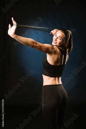 Young, slim girl doing exercises with rubber expander on black background.