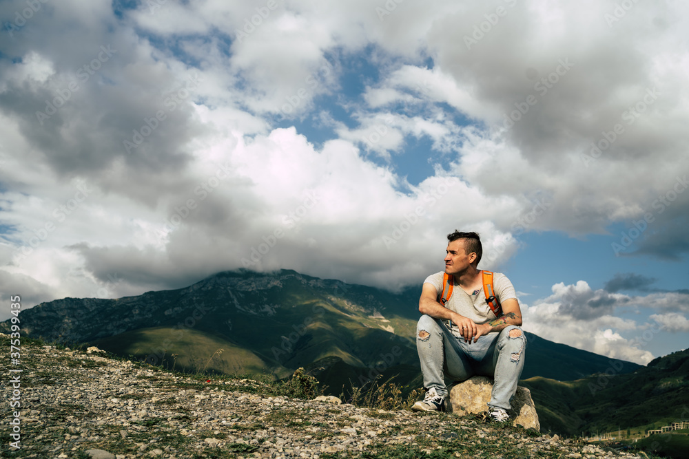 Young man sitting on hill. Dreamy male sitting on altitude and looking at Peaks of magnificent rocks located against bright cloudy sky