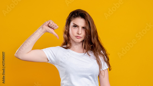 Dislike gesture. Bad idea. Portrait of dissatisfied unhappy woman in white t-shirt rejecting with thumb down isolated on orange copy space background. No sign. Negative reaction. Leave it.