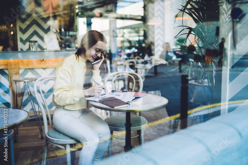 Young woman talking on phone and making notes in cozy cafe