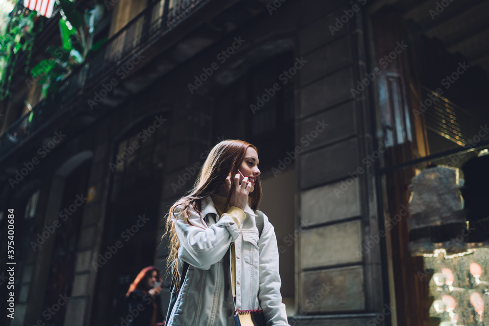 Fototapeta premium Joyful modern female student talking on smartphone while walking along street