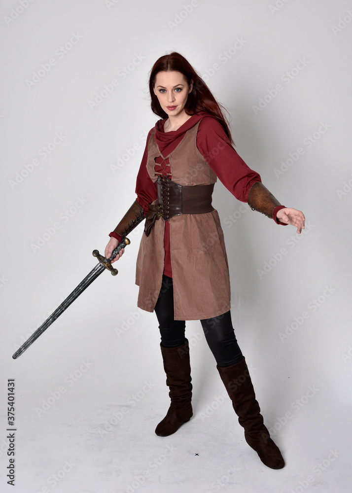 Full length portrait of girl wearing medieval costume. Standing pose holding a sword, isolated against a grey studio background. Full length portrait of girl wearing medieval costume. Standing pose holding a sword, isolated against a grey studio background.