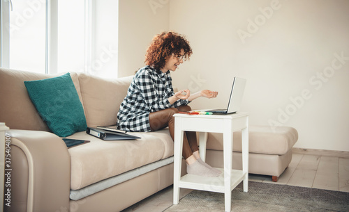 Side view photo of a caucasian lady with curly hair and eyeglasses smiling while having online lessons using a laptop at home