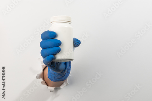 Hand in a latex glove holding a plastic bottle of pills through torn white wall
