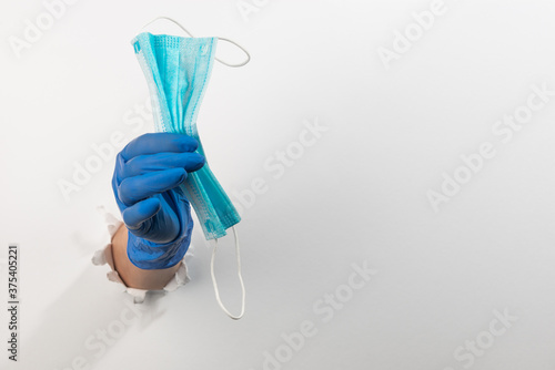 Hand in a latex glove giving a surgical face mask throught torn white wall