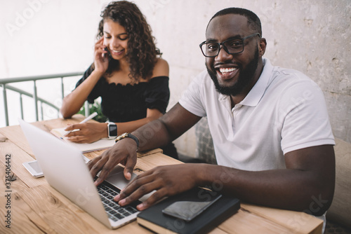 Portrait of dark skinned male freelancer in optical spectacles for provide eyes protection smiling at camera during collaboration meeting with successful woman, happy colleagues in coworking