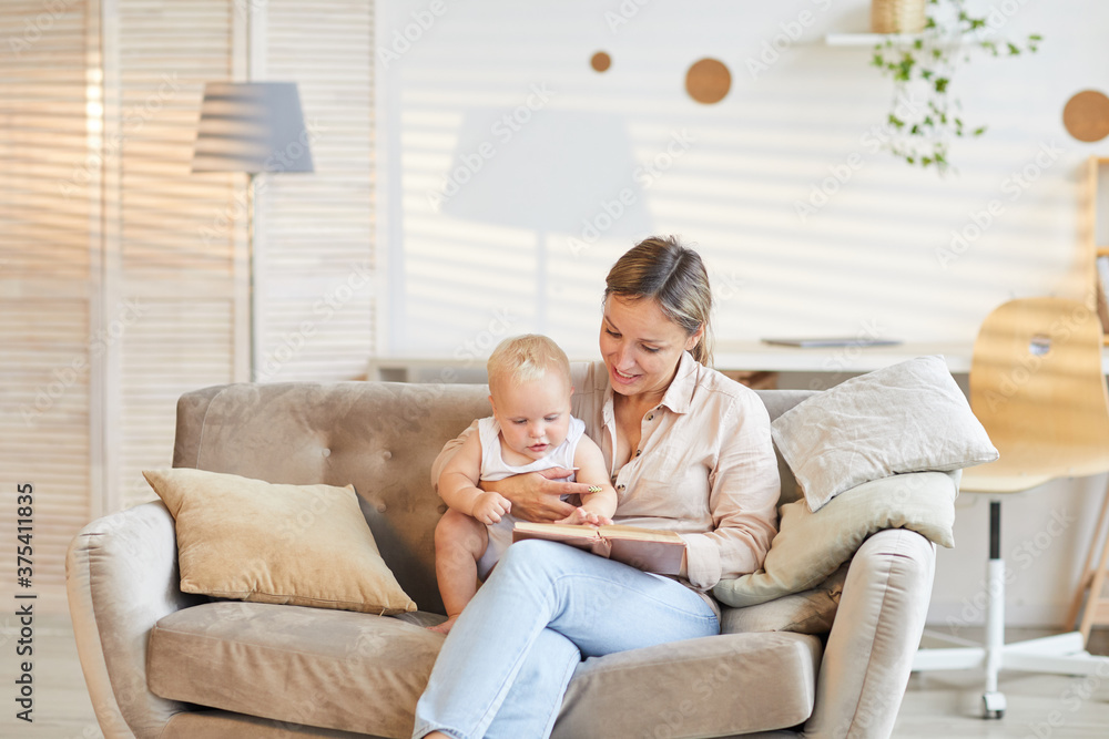 Modern young mother sitting on sofa in living room with her curious kid reading book, copy space