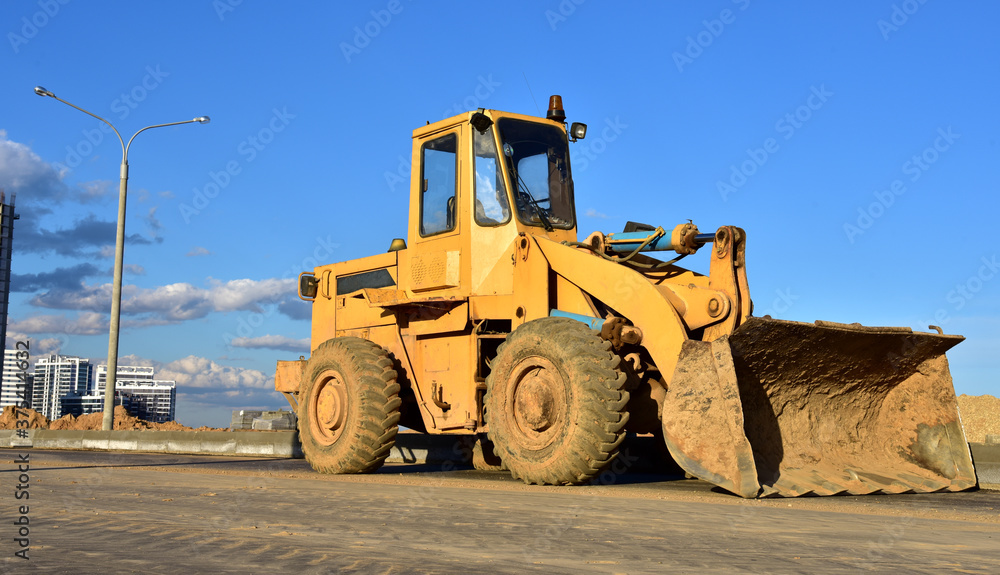 Wheel loader with a bucket on a street in the city during the construction of the road. Construction site with heavy machinery for road work. Public works, civil engineering, road building.
