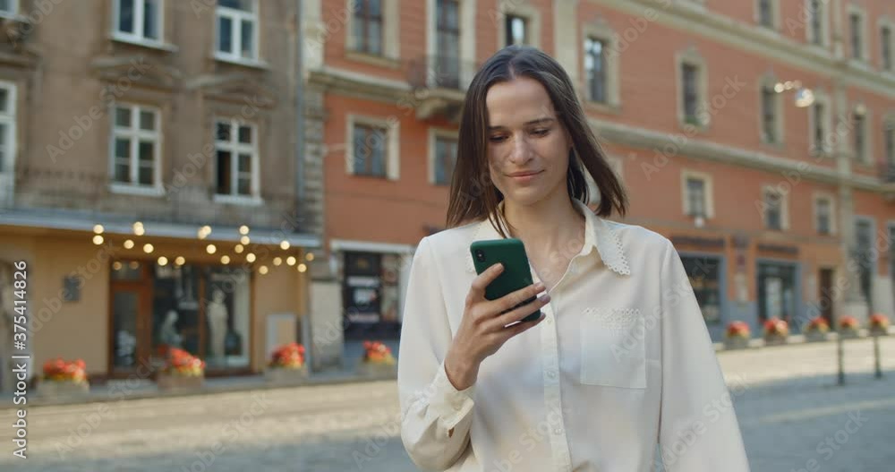 Portrait of female student girl looking at her mobilephone screen while walking at old city street. Beautiful young woman scrolling news feed and smiling while using phone outdoors. Portrait of female student girl looking at her mobilephone screen while walking at old city street. Beautiful young woman scrolling news feed and smiling while using phone outdoors.