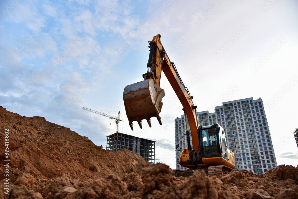 Excavator during earthmoving at construction site on sunset background. Сonstruction machinery for excavating. Tower cranes lifting a concrete bucket for pouring of concrete into formwork