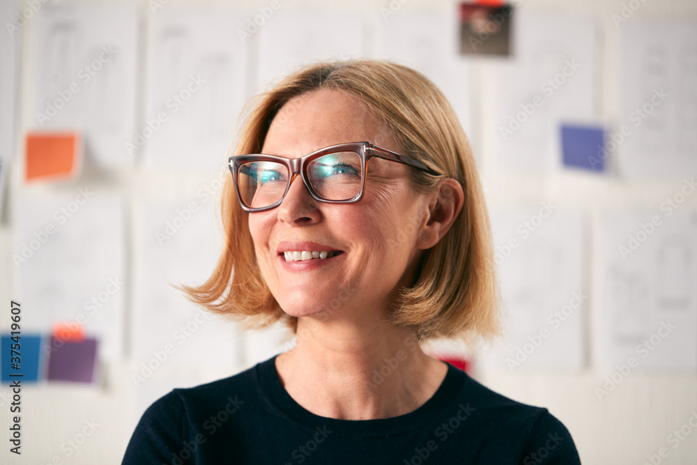 Portrait Of Mature Woman Standing In Front Of Designs On Wall In Start Up Fashion Business Portrait Of Mature Woman Standing In Front Of Designs On Wall In Start Up Fashion Business