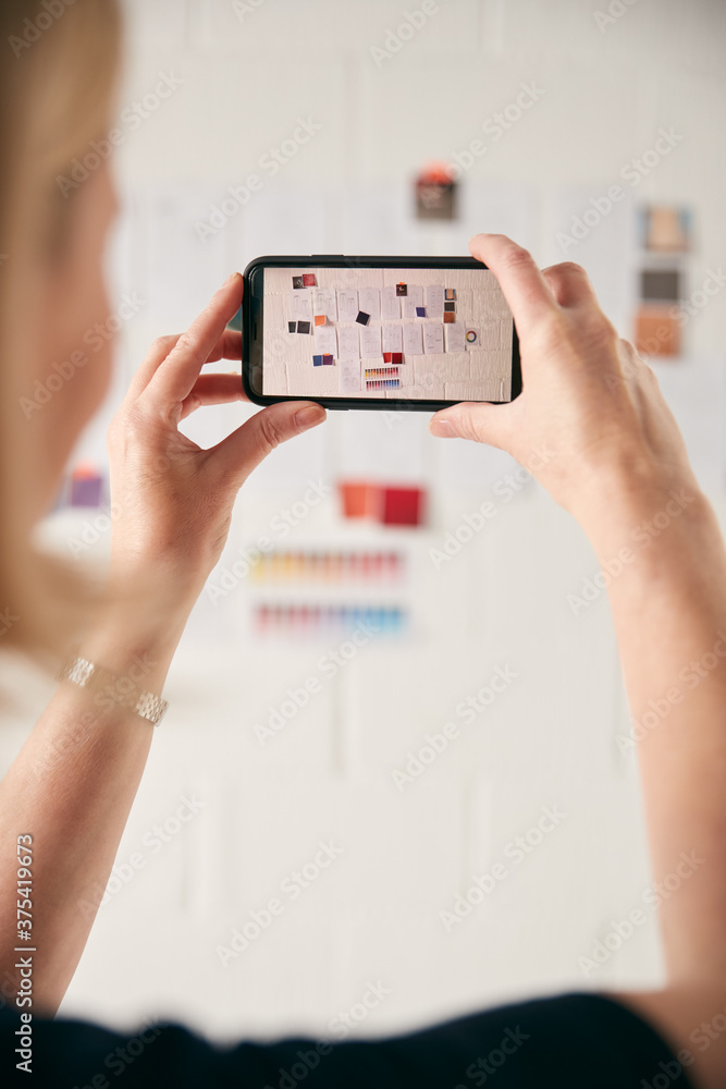 Female Designer Taking Photos Of Designs On Wall With Mobile Phone In Start Up Fashion Business Female Designer Taking Photos Of Designs On Wall With Mobile Phone In Start Up Fashion Business