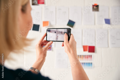 Female Designer Taking Photos Of Designs On Wall With Mobile Phone In Start Up Fashion Business