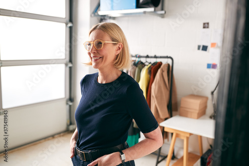 Portrait Of Mature Businesswoman Standing In Front Of Designs And Desk In Start Up Fashion Business