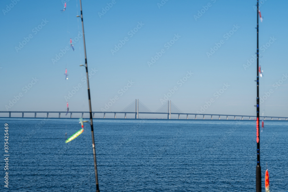 Fishing rods with the Oresund Bridge linking Denmark with Sweden in the background. Clear blue sky and a calm blue ocean