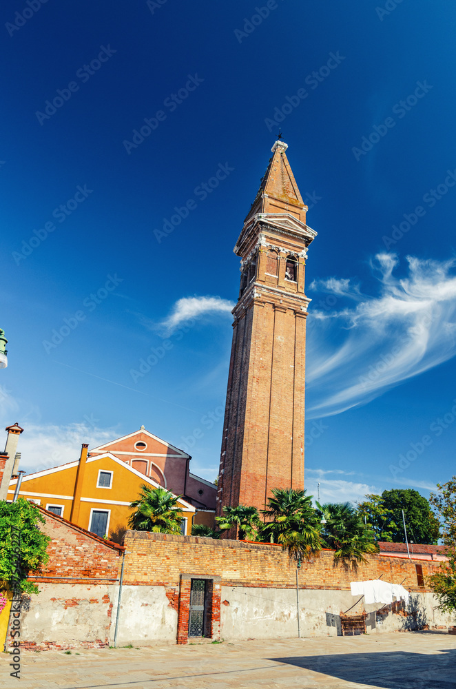 Obraz premium San Martino Roman Catholic church in Burano island with brick bell tower campanile, blue sky background in sunny summer day, Venice Province, Veneto Region, Northern Italy. Burano postcard