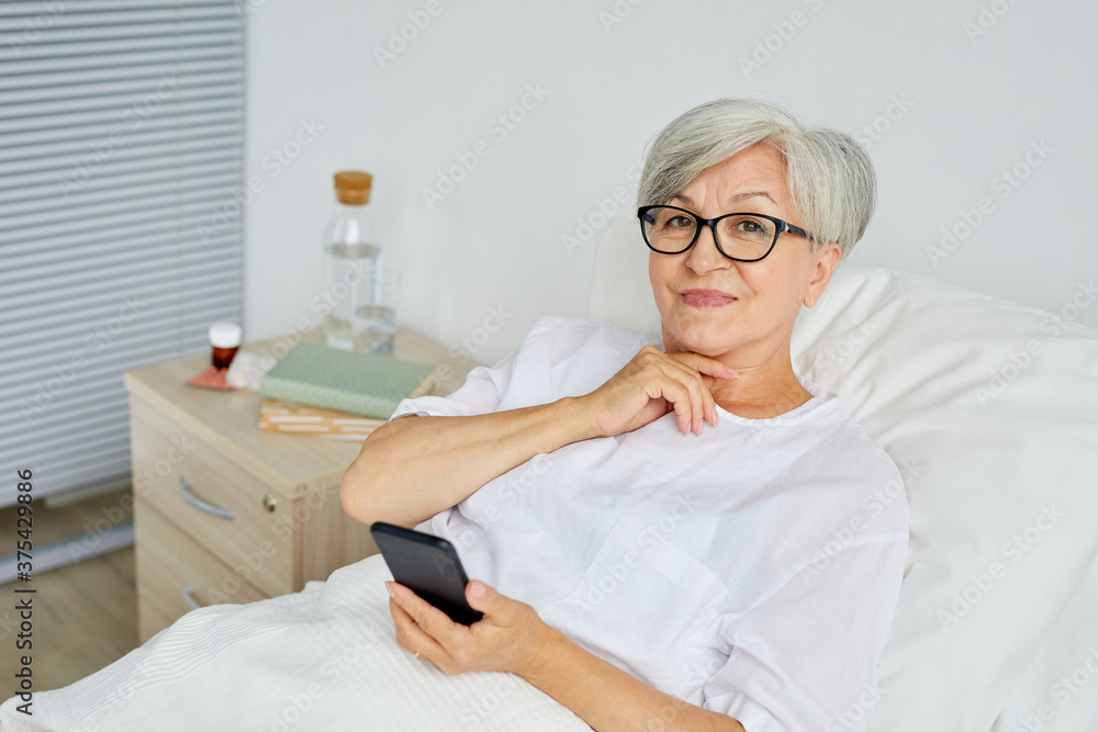 Fototapeta premium Portrait of beautiful aged woman wearing white clothes lying on bed in hospital ward holding smartphone looking at camera