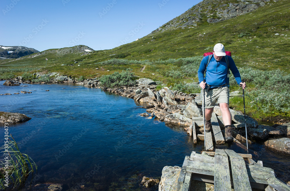 Hiking in Swedish Lapland. Man crossing river on small wooden broken bridge, Trekking alone in north Sweden. Arctic nature of Scandinavia in warm summer sunny day with blue sky