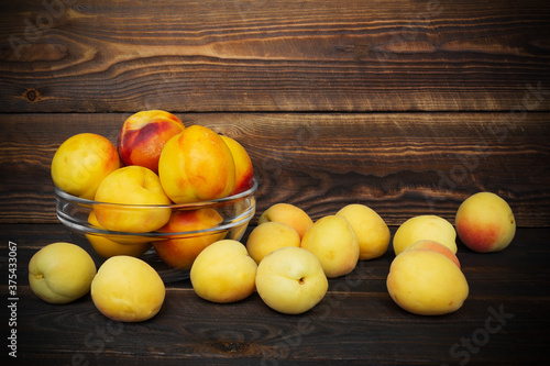 Nectarines and apricots in transparent glass bowl on brown wooden plank background, scattered fruits around