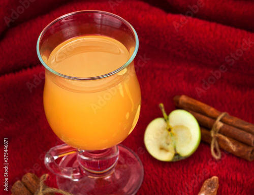 Hot apple drink in the glass mug with apple fruits and spices on a red textile background.