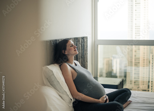 happy pensative pregnant woman sitting on bed (ID: 375449278)