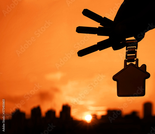 Buying or renting resindental house concept. Man hand holding 3 three silver key with houme shape keyring, on sunlight sky cityscapes background. photo