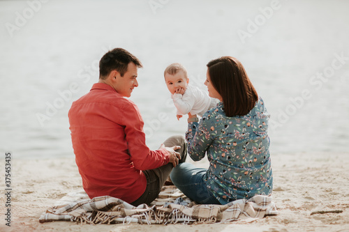 Happy young family sitting near the water. Mom, dad and their little baby daughter sitting together. Happy family outdoors. Smiling people.