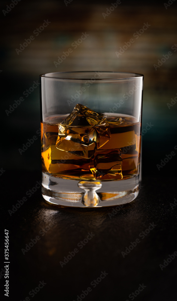 Glass of Kentucky Bourbon on rocks reflecting on black stone surface and refracting colorful wooden background. Glass of Kentucky Bourbon on rocks reflecting on black stone surface and refracting colorful wooden background.