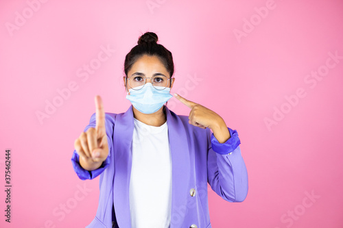 Young beautiful business woman over isolated pink background pointing the mask. Warning expression with negative and serious gesture on the face.