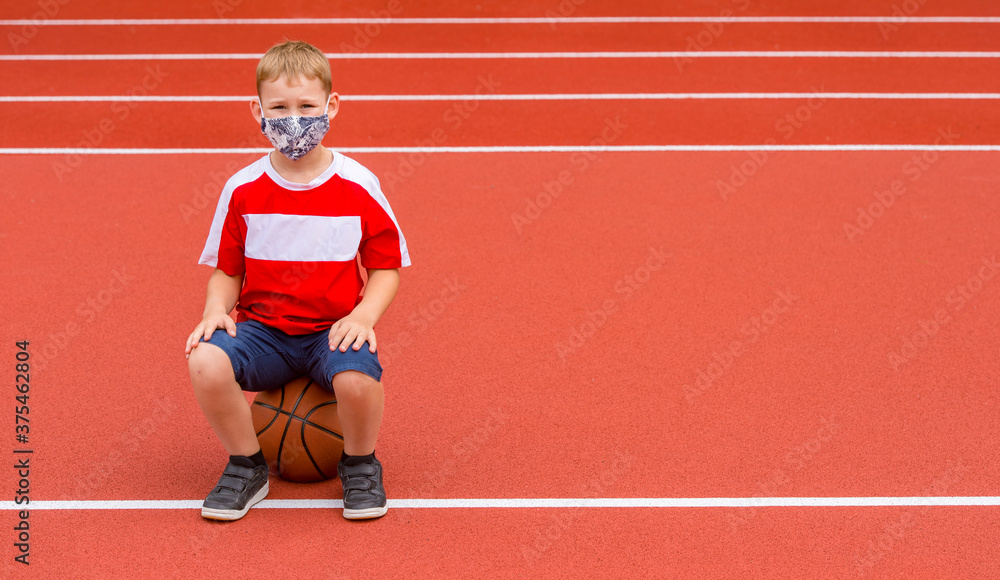 Obraz premium School kid with mask and basketball ball in a physical education lesson. Safe back to school during pandemic concept. Social distancing to fight COVID-19