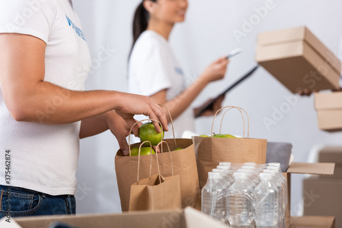 Cropped view of volunteer putting apples in paper bags near bottles of water in charity center