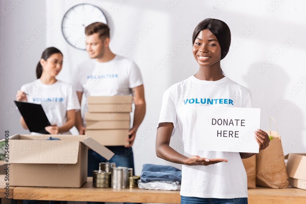 Selective focus of african american volunteer pointing with hand at card with donate here lettering in charity center Selective focus of african american volunteer pointing with hand at card with donate here lettering in charity center