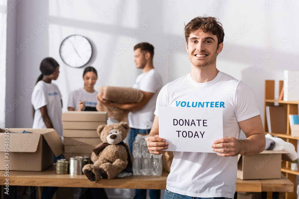 Obraz premium Selective focus of volunteer with donate today lettering on card looking at camera near multiethnic people in charity center