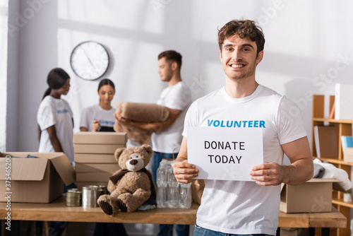 Selective focus of volunteer with donate today lettering on card looking at camera near multiethnic people in charity center