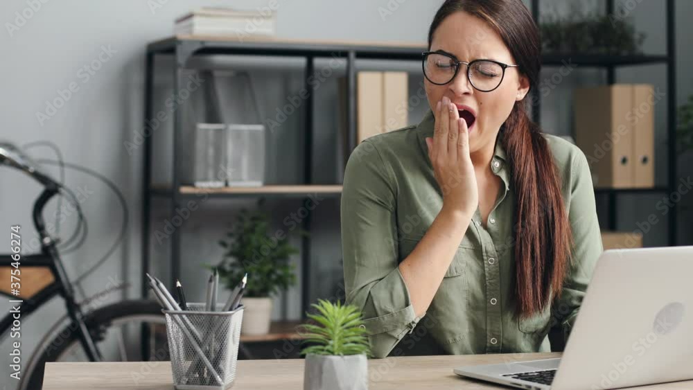 Young beautiful woman with glasses working on laptop. Female worker yawns while sitting at her desk in modern office. Young beautiful woman with glasses working on laptop. Female worker yawns while sitting at her desk in modern office.
