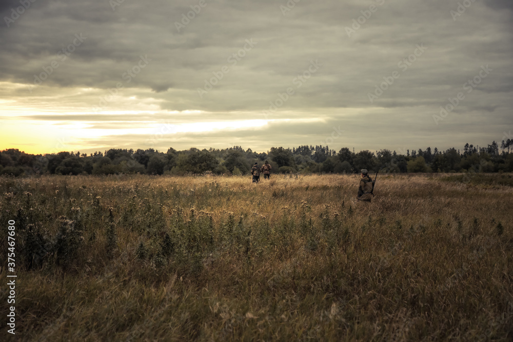 Hunters men during hunting standing in rural field and looking into the distance forest at beautiful sunset as hunting lifestyle