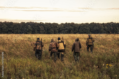 Hunters going through rural field towards forest against sunrise sky and forest on horizon during hunting season