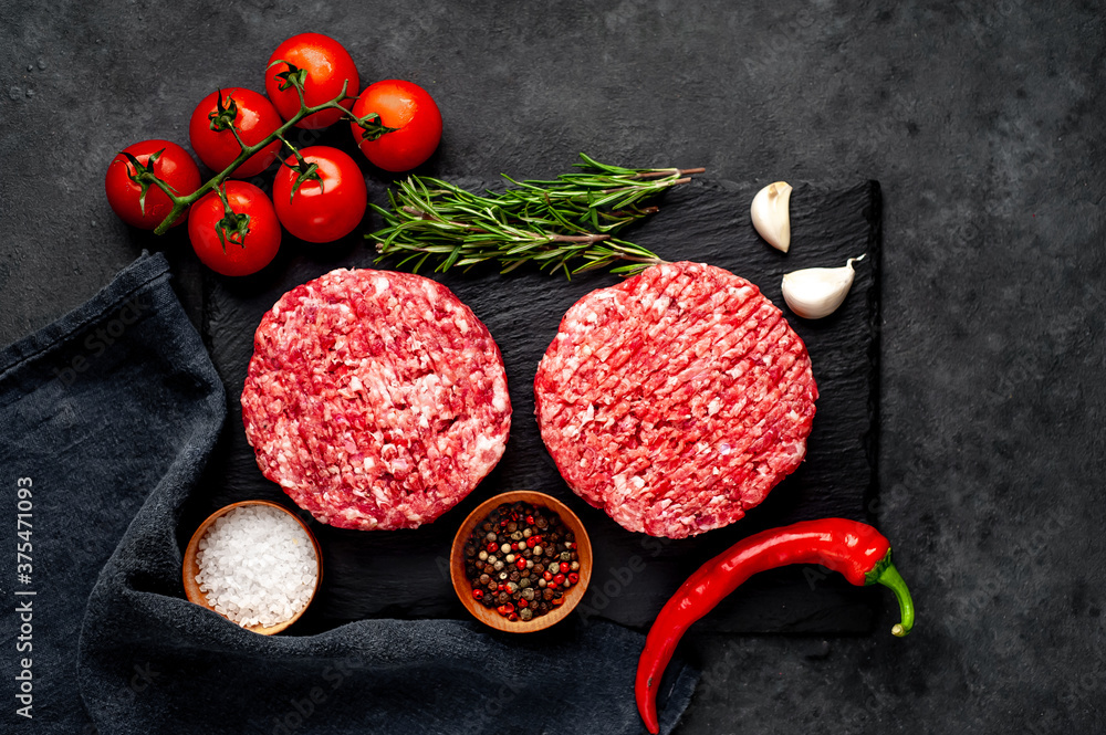 Raw beef burger cutlets with rosemary and spices on a slate board on a concrete background