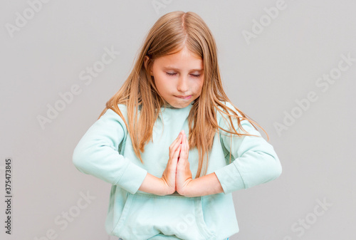 Namaste, friends. Portrait of friendly-looking happy european female student in casual clothes, holding palms in pray and smiling broadly while making buddhist greeting or praying over gray wall
