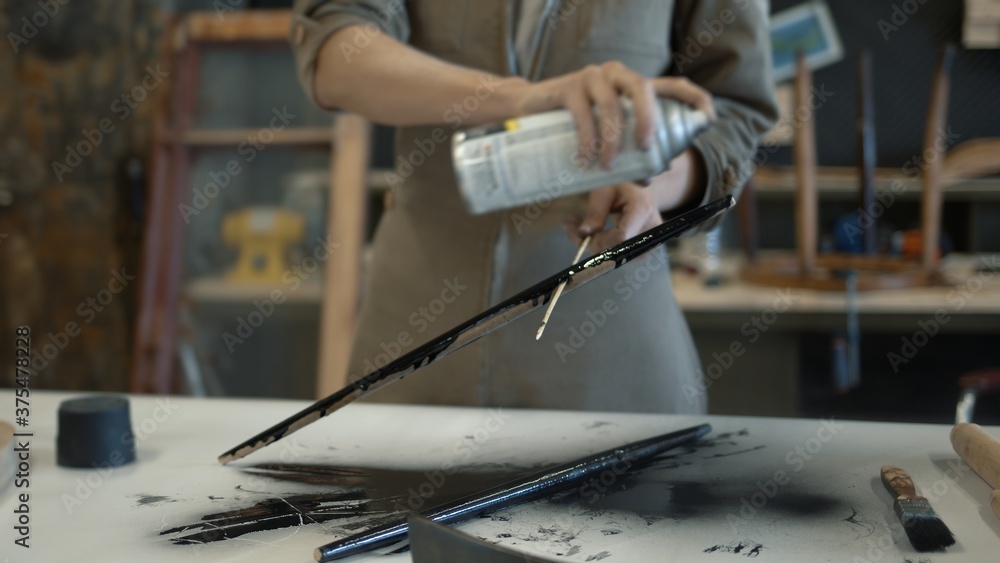 Fototapeta premium A female restorer spraying a wooden detail with black color can, female hands renew old wooden furniture. Carpenter working with wooden material in workshop