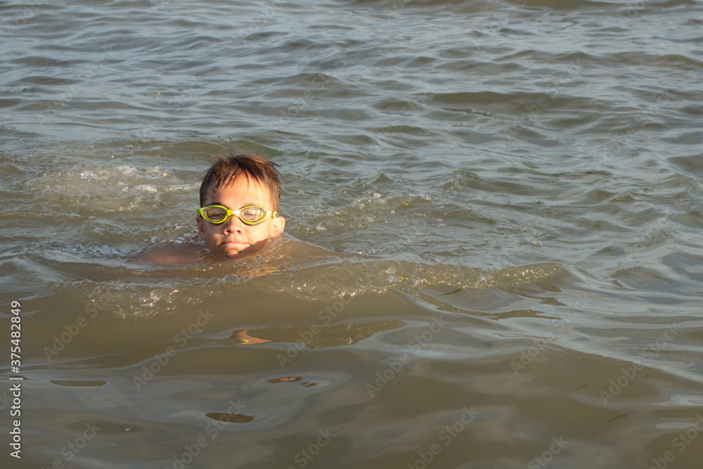 A 10-year-old boy in green swimming goggles swims in the sea near the shore.