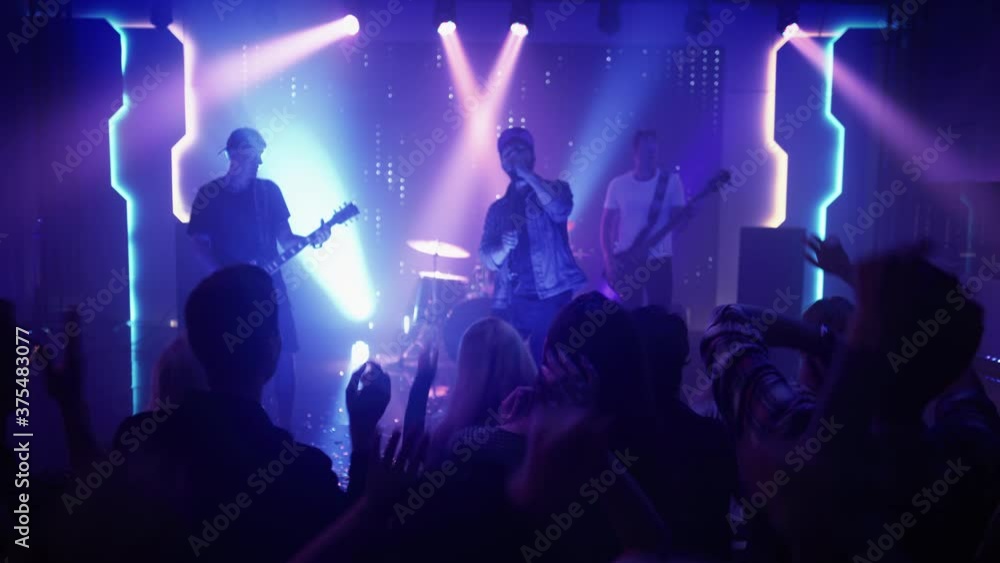 Rock Band with Guitarists and Drummer Performing at a Concert in a Night Club. Front Row Crowd is Partying. Silhouettes of Fans Raise Hands in Front of Bright Colorful Strobing Lights on Stage. Rock Band with Guitarists and Drummer Performing at a Concert in a Night Club. Front Row Crowd is Partying. Silhouettes of Fans Raise Hands in Front of Bright Colorful Strobing Lights on Stage.
