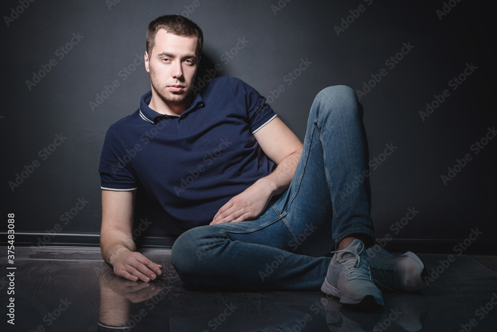 Young attractive man in blue jeans and a polo shirt sits on the floor near the wall in the studio. Youth and attractiveness concept