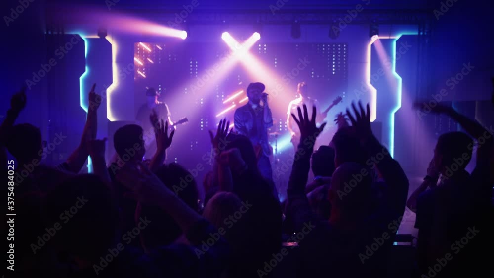 Rock Band with Guitarists and Drummer Performing at a Concert in a Night Club. Front Row Crowd is Partying. Silhouettes of Fans Raise Hands in Front of Bright Colorful Strobing Lights on Stage. Rock Band with Guitarists and Drummer Performing at a Concert in a Night Club. Front Row Crowd is Partying. Silhouettes of Fans Raise Hands in Front of Bright Colorful Strobing Lights on Stage.
