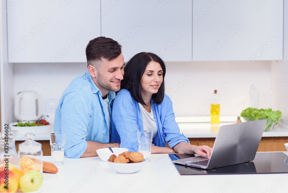 Beautiful happy young couple having breakfast at home and uses laptop at kitchen. Enjoying free time together. Beautiful happy young couple having breakfast at home and uses laptop at kitchen. Enjoying free time together.