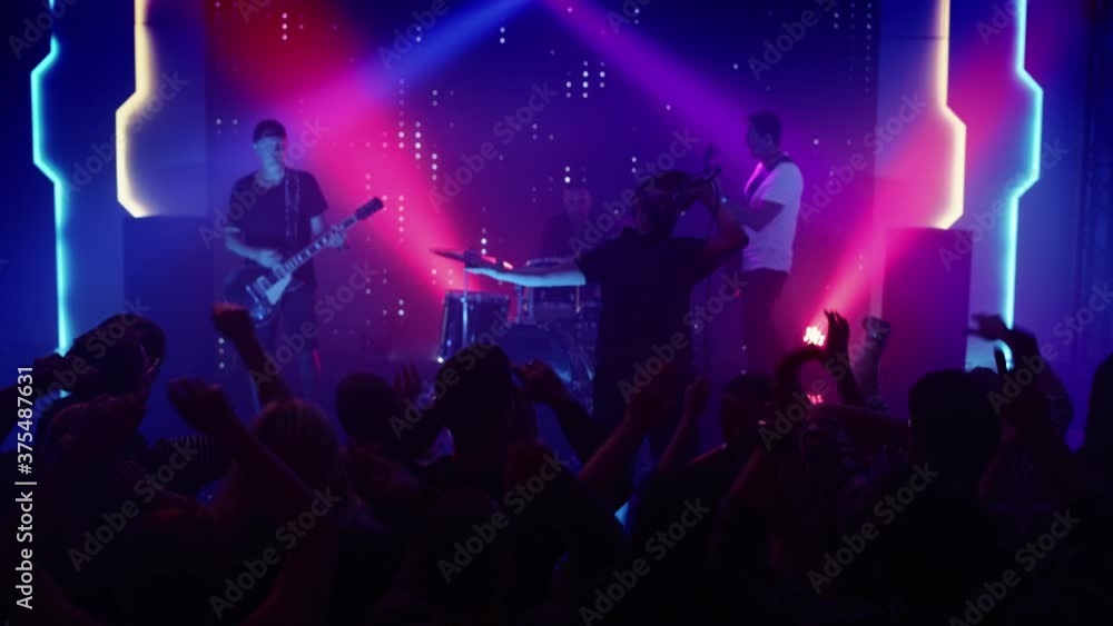 Rock Band with Guitarists and Drummer Performing at a Concert in a Night Club. Front Row Crowd is Partying. Silhouettes of Fans Raise Hands in Front of Bright Colorful Strobing Lights on Stage. Rock Band with Guitarists and Drummer Performing at a Concert in a Night Club. Front Row Crowd is Partying. Silhouettes of Fans Raise Hands in Front of Bright Colorful Strobing Lights on Stage.