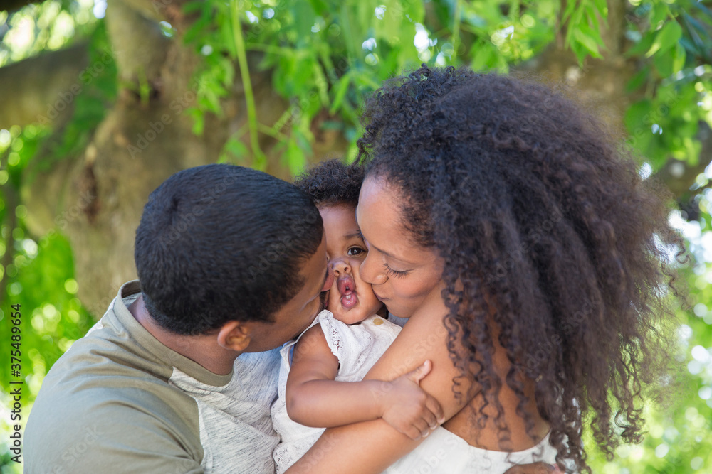 Affectionate parents kissing toddler daughter below tree Affectionate parents kissing toddler daughter below tree