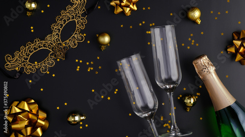 Champagne bottle with confetti stars, glasses, golden holiday decorations, masquerade mask on black background. Christmas party or New Year celebration concept. Flat lay, top view.